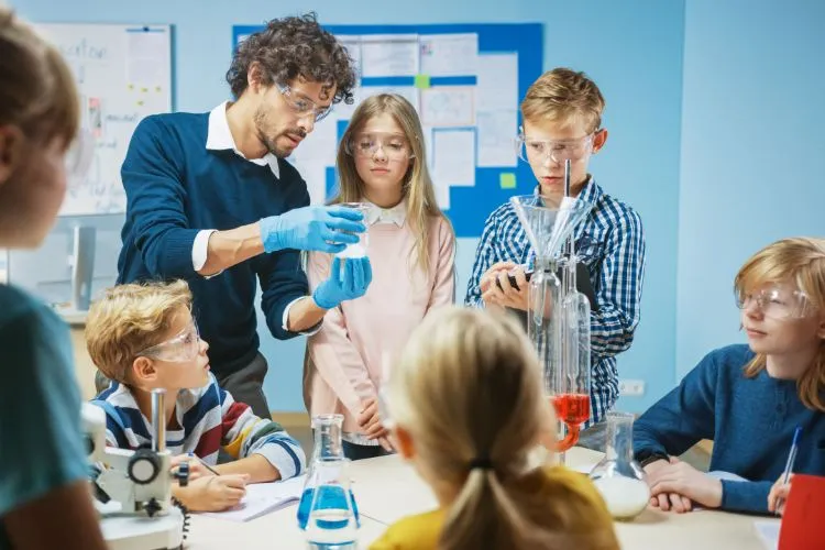 science-teacher-teaching-young-school-students-in-a-classroom.jpg A science teacher teaches five schoolchildren in a classroom, while wearing safety goggles and doing something with glassware