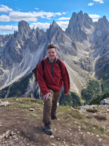 Robert Dryfe wearing a red fleece pullover and a backpack stands on a dirt path in front of a rugged mountain range.