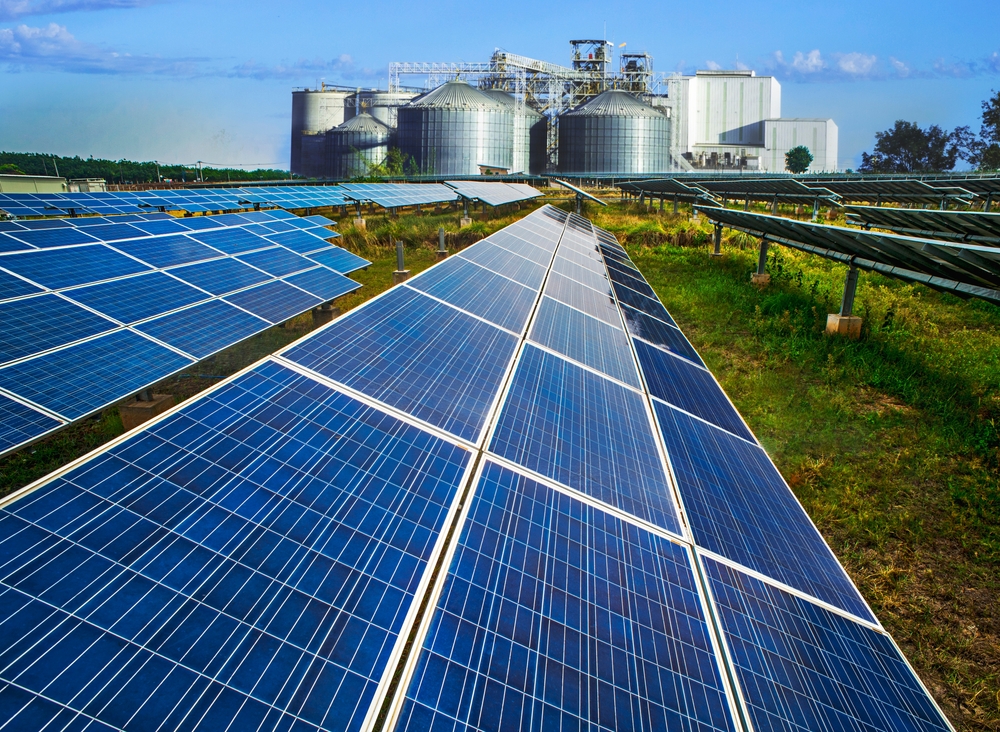 A large bank of solar panels in the foreground with an industrial plant in the background