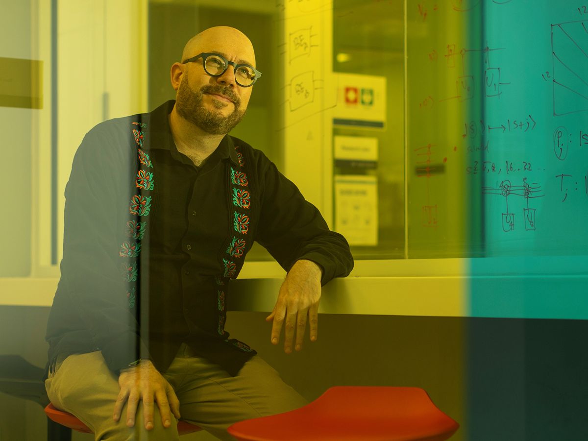 Alán Aspuru-Guzik sitting on stool leaning on shelf in lecture lab, wearing glasses and black shirt with coloured patterned squares