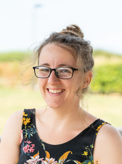 Heather Fish smiling, wearing black rectangular glasses and a floral print sleeveless top.