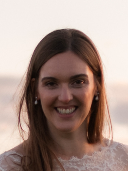 Emma Jordan-Rose smiling broadly, wearing a beige lace top and out-of-focus view of a beach in the background.