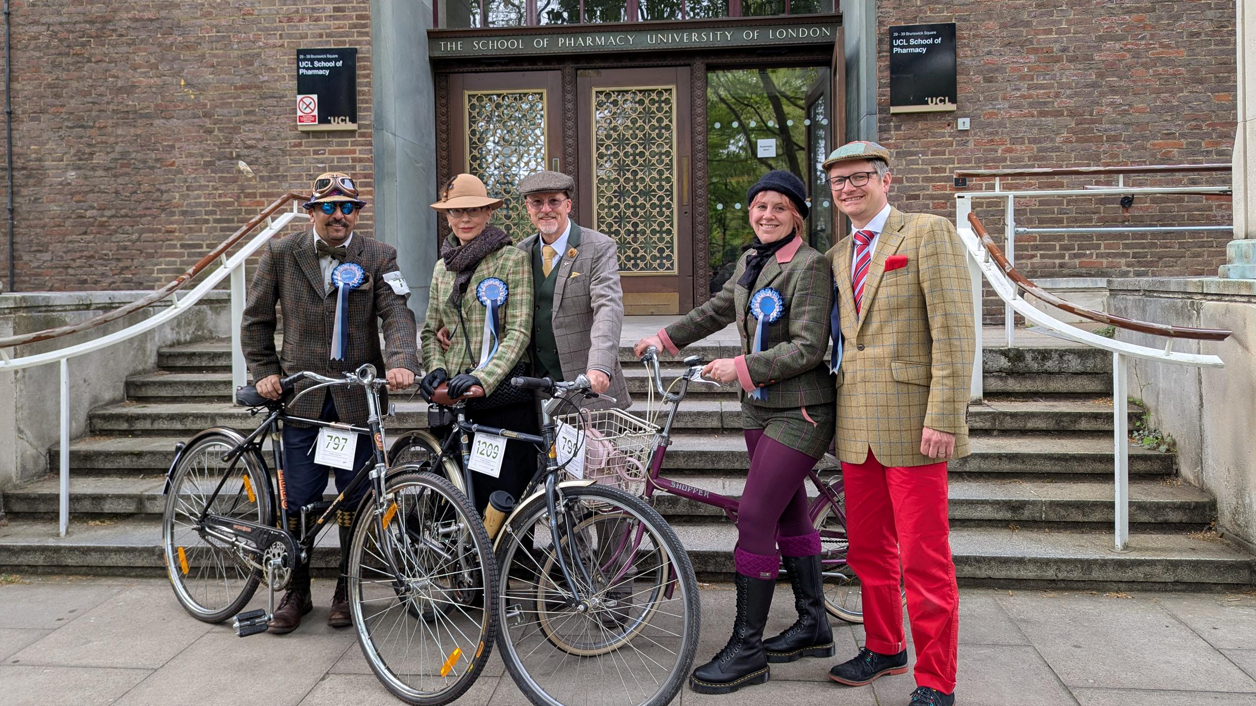 Zoe with others and their bikes, wearing suits and hats standing in front of a building