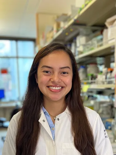 Miranda Villaneuva Miranda Villaneuva smiling in a white laboratory coat, posed in a science research lab with equipment shelves visible in the background.