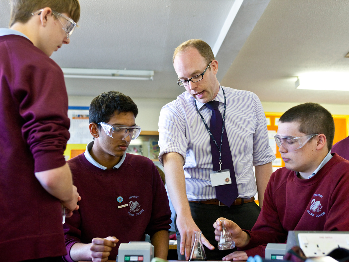Three male school pupils in their early teens, wearing safety goggles and burgundy jumpers sit either side of a tall science teacher, who is wearing glasses, a shirt and tie and a lanyard