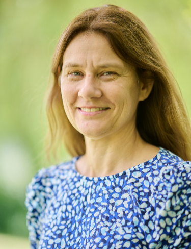Lynette Holland smiling, wearing a blue and white patterned blouse, set against a soft, blurred natural green background.