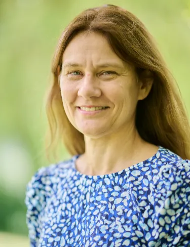 Lynette Holland Lynette Holland smiling, wearing a blue and white patterned blouse, set against a soft, blurred natural green background.