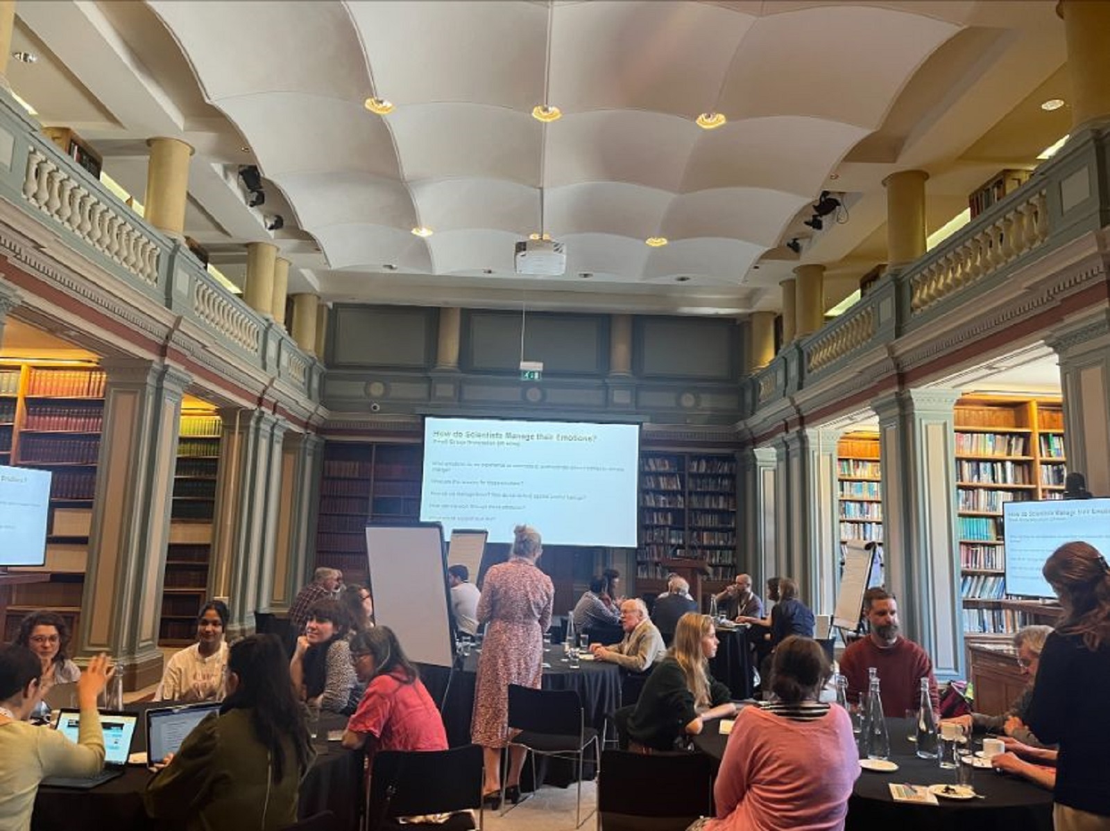 A landscape image of a grand room in Burlington House with books along the walls and event attendees sat around circular tables in discussion with a screen asking 'How do scientists manage their emoti
