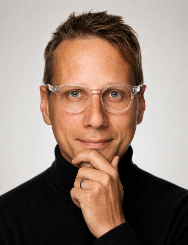 Mathias Winkel wearing a black turtleneck and clear glasses, posed with his hand resting on his chin against a white background.