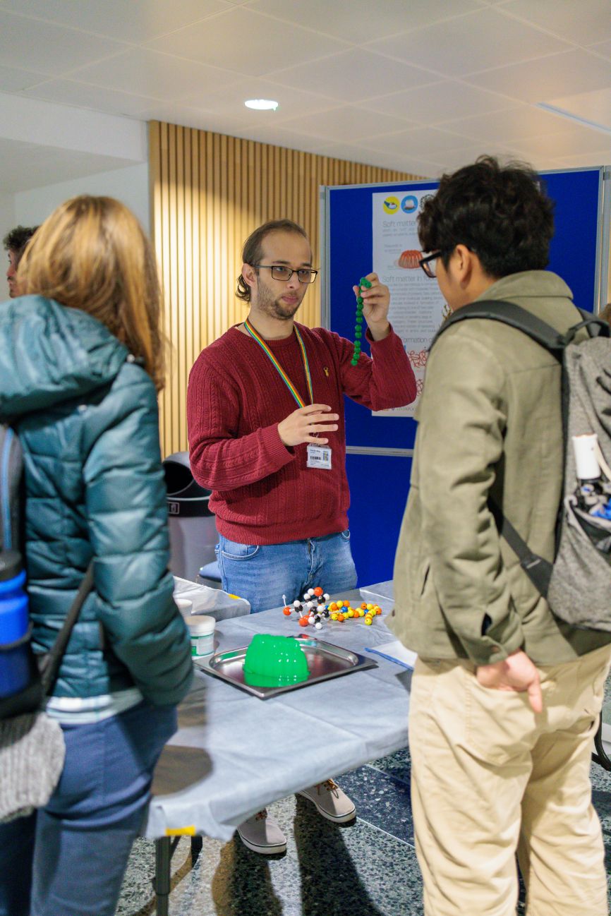 A man in glasses and a red jumper talks to two people at a stall at the Imperial Lates event