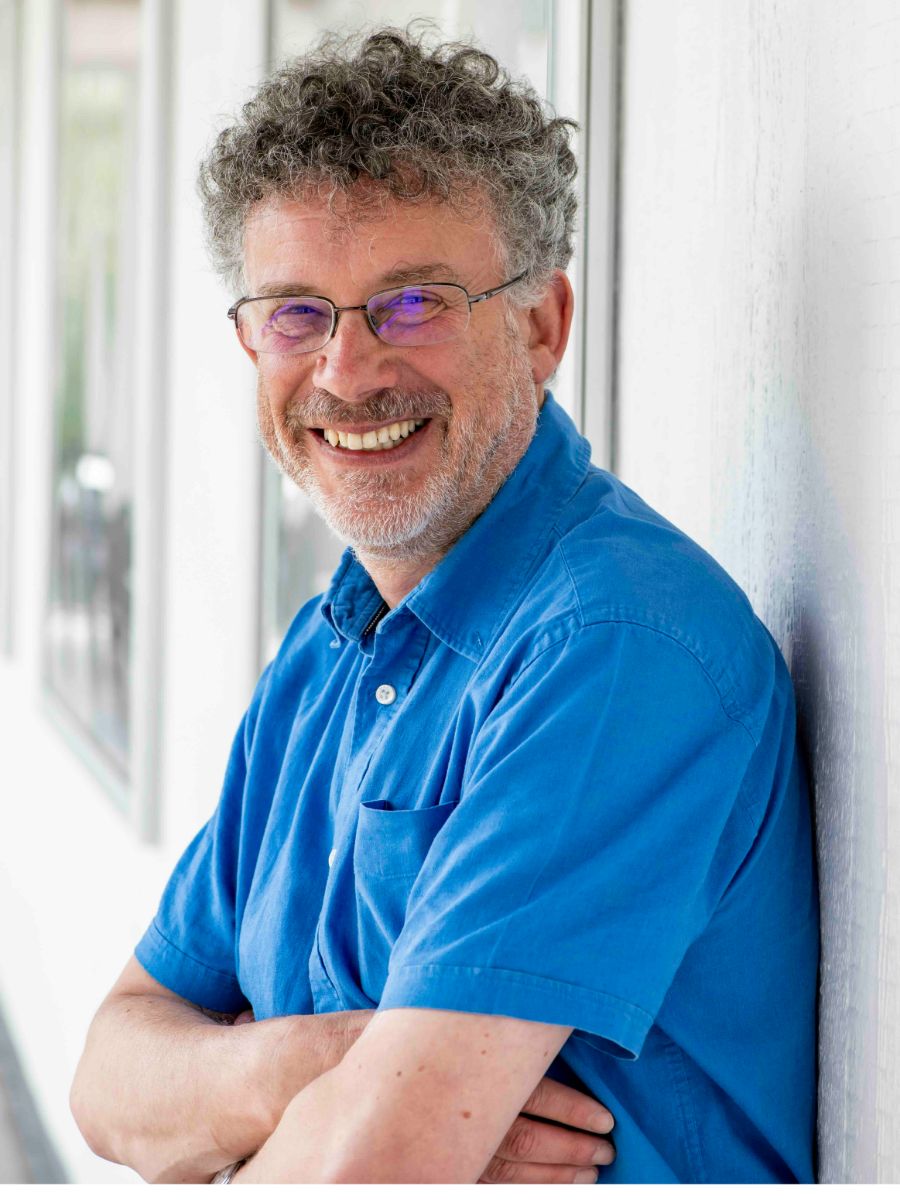Jeremy Frey smiling at camera leaning against a white wall, wearing glasses and a blue shirt