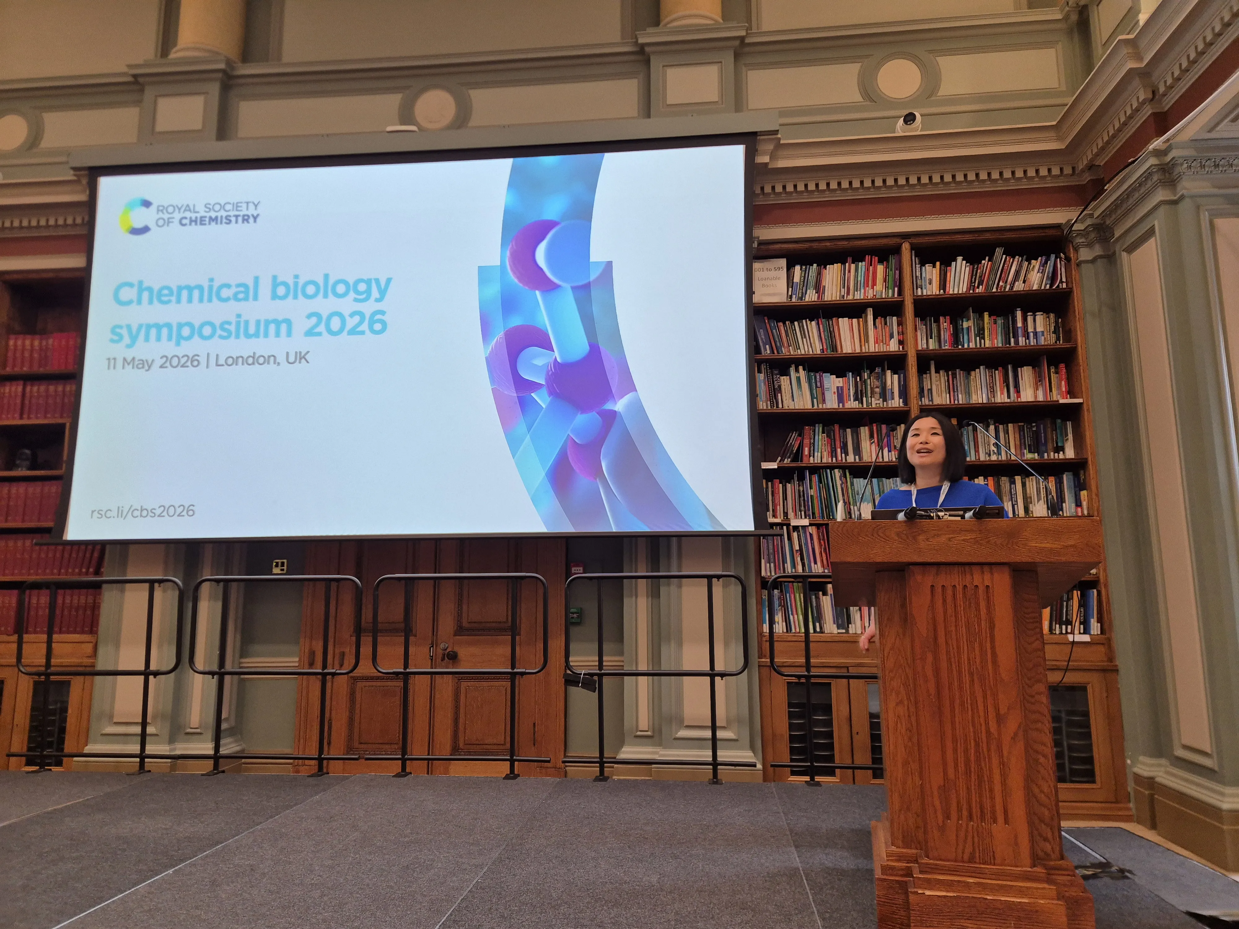 2026 Chemical Biology Symposium - Akane Kawamura opening remarks Professor Akane Kawamura stands behind a lectern to the right of the image, with a screen reading Chemical Biology Symposium 2026 on the other side of the imagerge screen