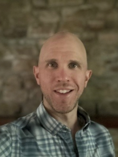 M Kevin Brown smiling at the camera, wearing a blue and grey shirt against a blurred stone wall background.