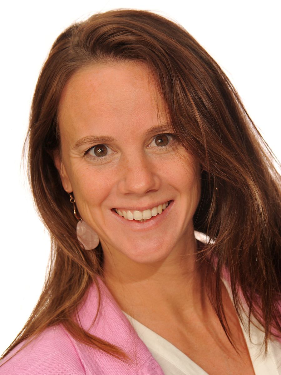 Elke Debroye headshot, smiling to camera with long brown hair and baby pink jacket