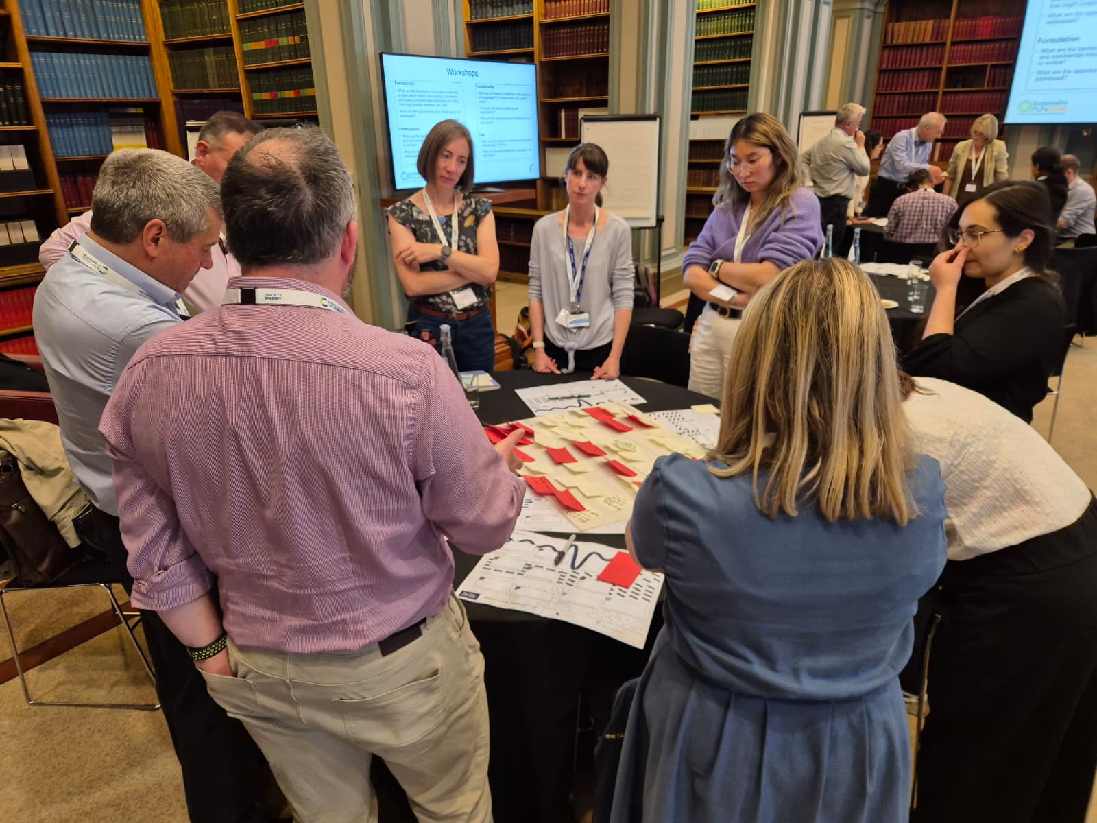 A group of nine people stand and talk while stood around a circular table with a big piece of paper in the middle covered in Post-it notes.