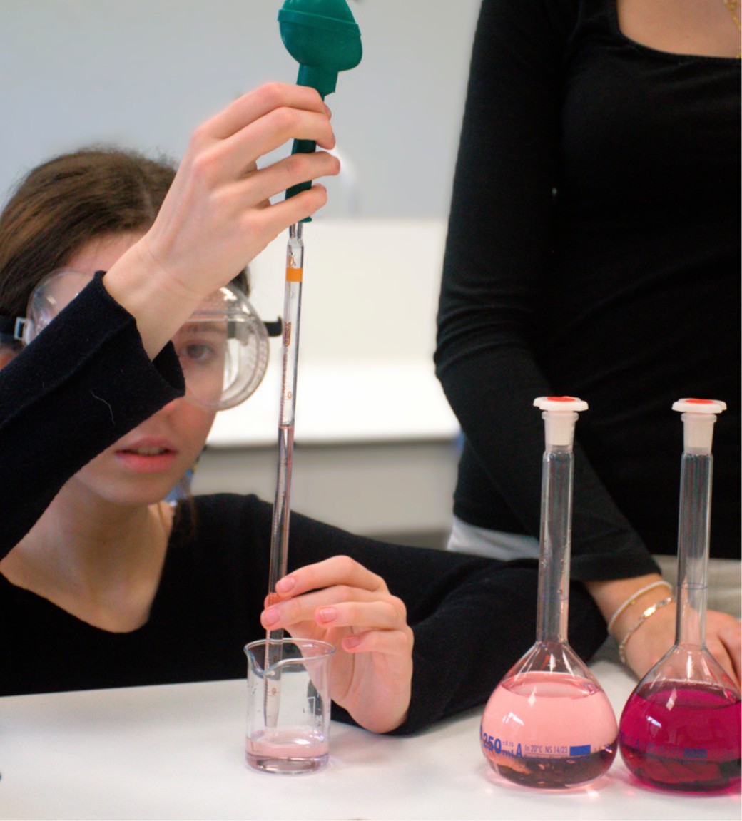 A young school girl in lab wearing safety goggles doing a science experiment using a pipette and beaker