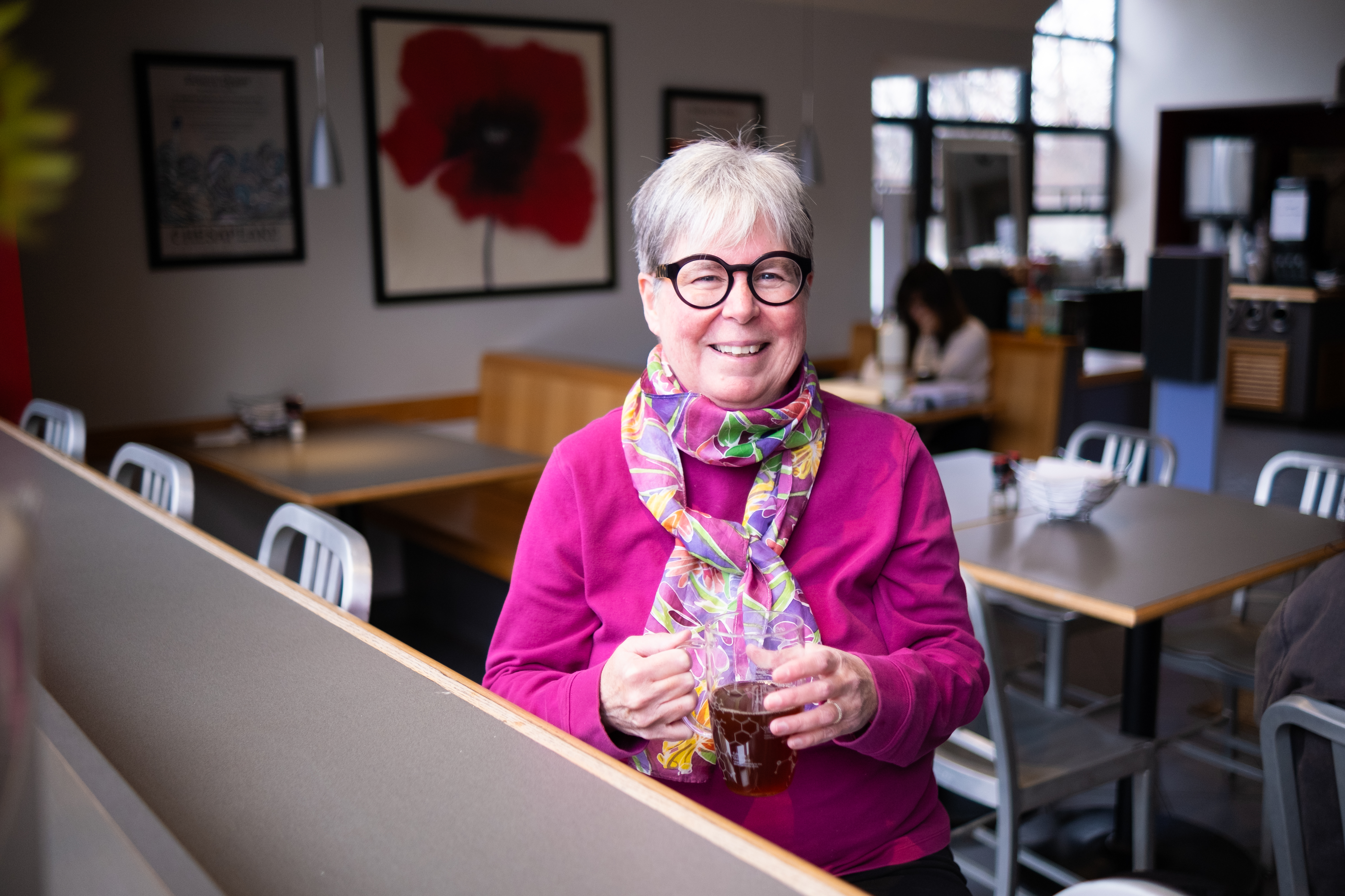 A smiling Dr Michelle Francl holds a glass cup of tea at an empty cafe