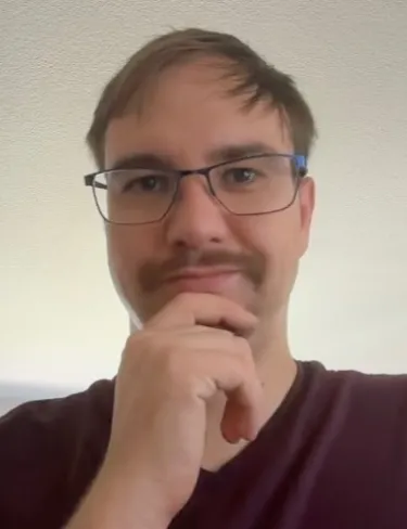 Alexander Hopp Alexander Hopp with light brown hair, glasses, and a mustache posing thoughtfully with his hand on his chin.