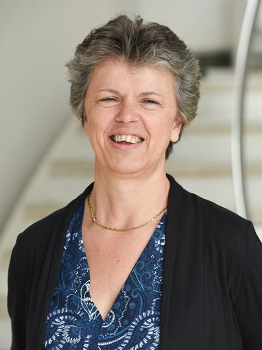 A head-and-shoulders photograph of a smiling Professor Gill Reid, wearing a black jacket and a blue top, in front of a whitewashed background with some stairs