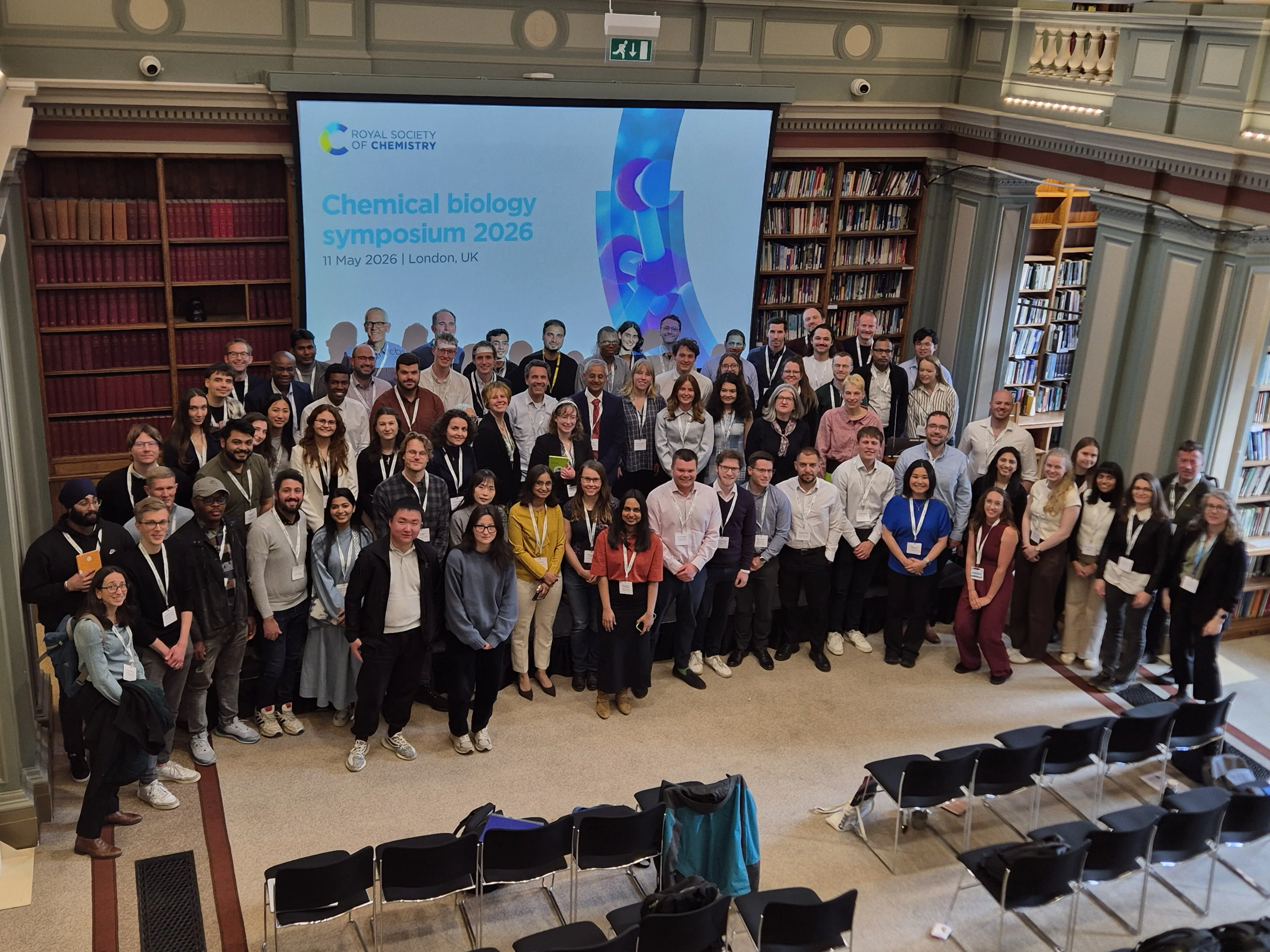 2026 Chemical Biology Symposium - delegates pose on stage Dozens of delegates stand in a room with a large screen