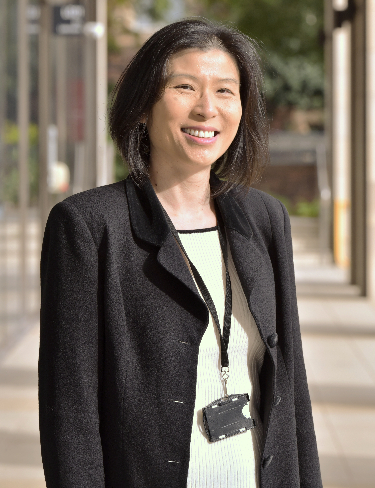  Anita Ho-Baillie smiling as she stands outdoors, wearing a dark jacket over a white top, and a lanyard with a badge holder hangs around her neck. 