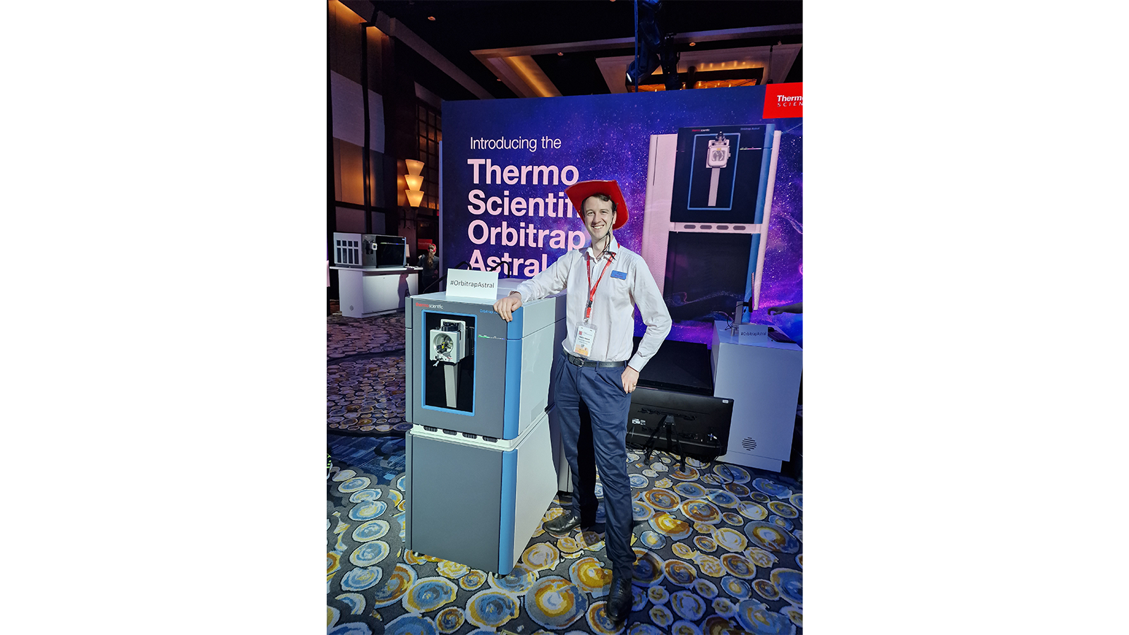 Orbitrap Astral Team member standing next to a large machine at an event, wearing a red cowboy hat!