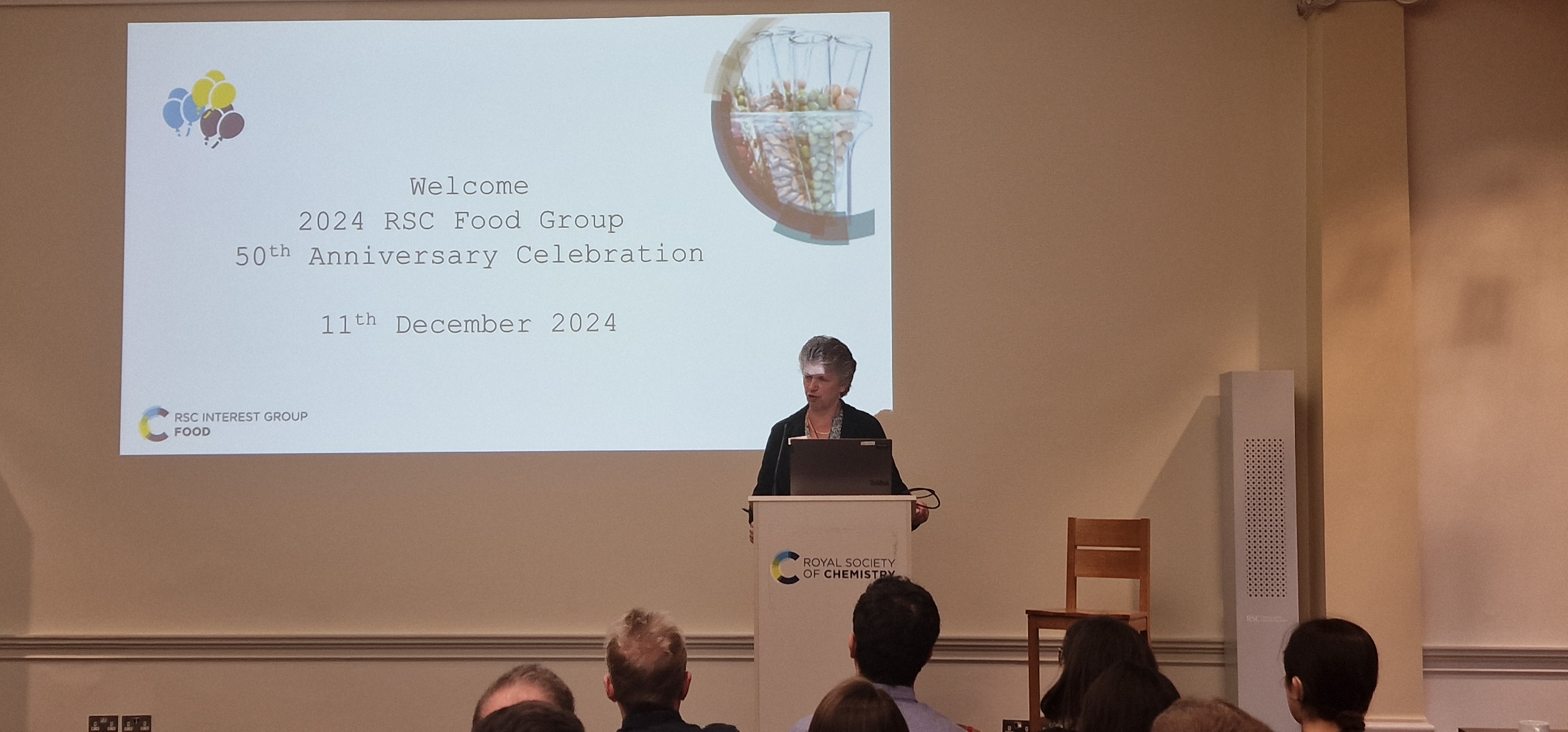 Former RSC President Professor Gill Reid stands behind a lectern and delivers a speech to the group, with the backs of five or six heads visible in the photo. She is stood in front of a large screen.