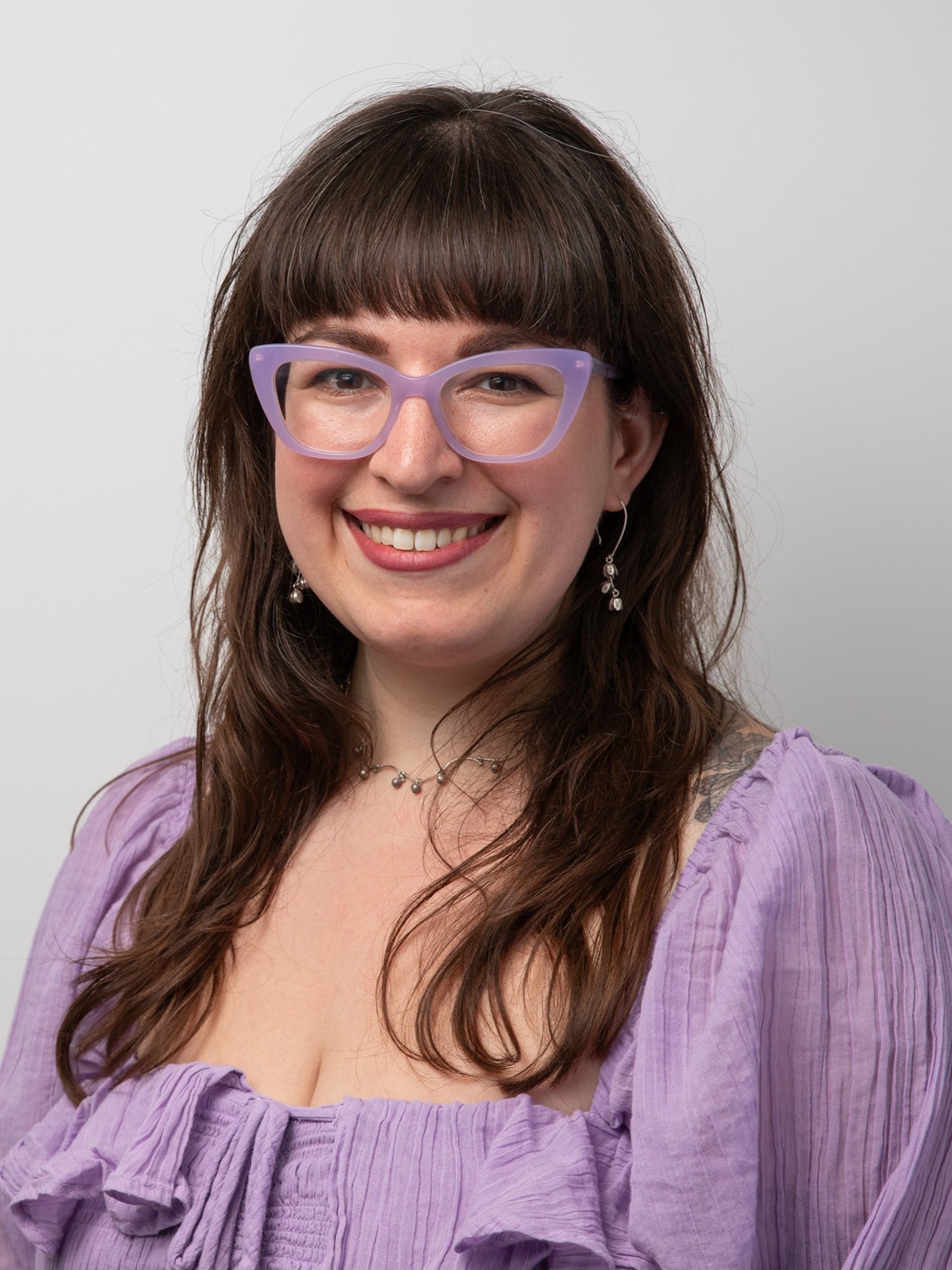 Sarah Holmes smiling, wearing a light purple top with a matching purple glasses, set plain grey background.