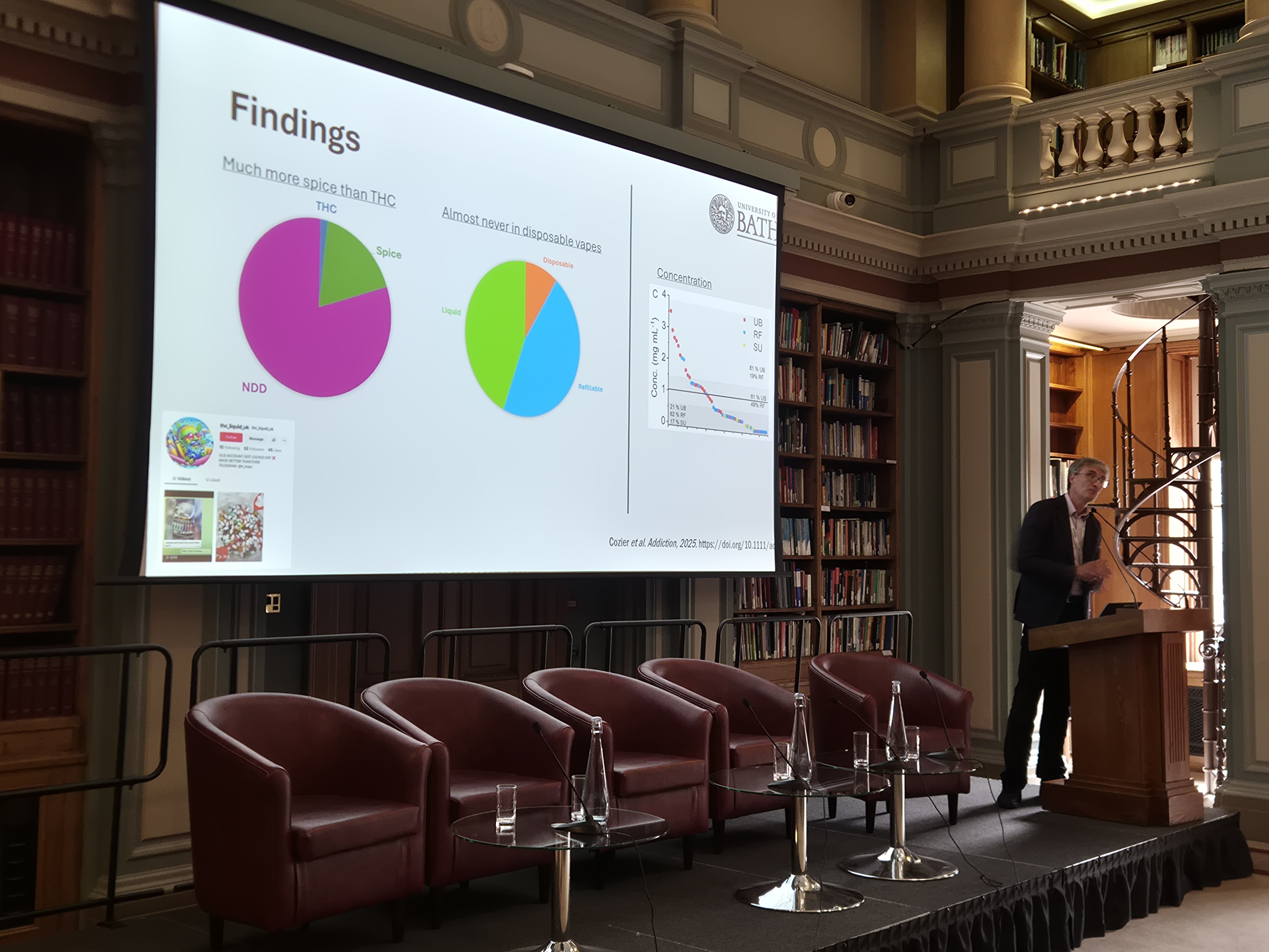 Stephen Husbands presents from behind a lectern while a big screen beside him shows a pair of pie charts