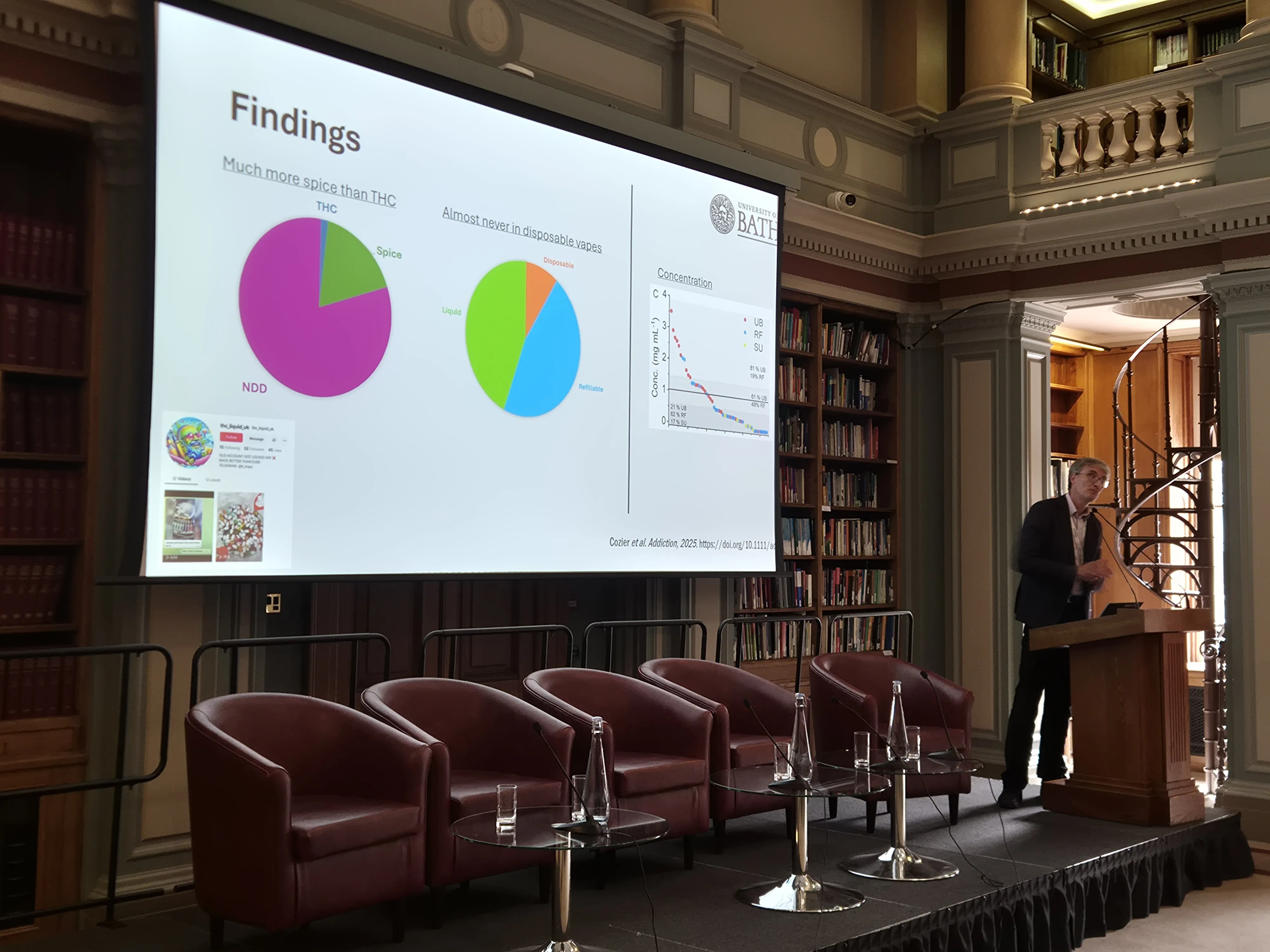 Analytical Research Forum 2026 - Stephen Husbands on spice in vapes Stephen Husbands presents from behind a lectern while a big screen beside him shows a pair of pie charts