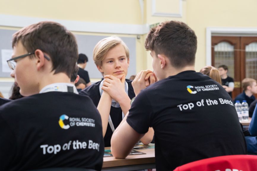 A student thinks about a question while two others, facing away from the camera wear Top of the Bench shirts, during the Top of the Bench final