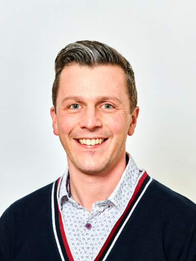 Stephen Cox smiling, wearing a patterned shirt underneath a navy blue sweater, set against a plain light background.