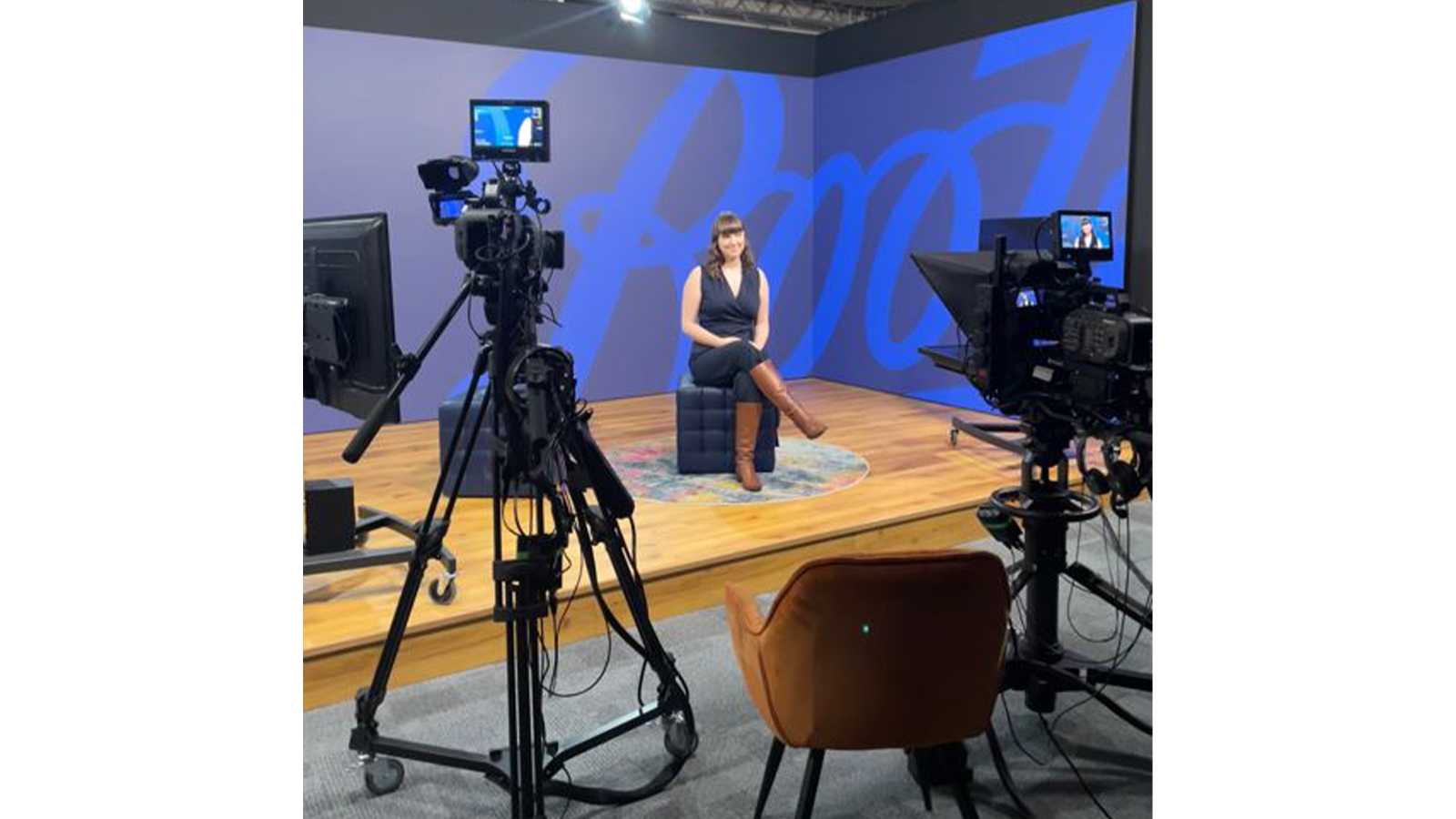 Mia Outram sitting on a chair in front of a blue wall with Boots the Chemist written on it, in front of many cameras in studio