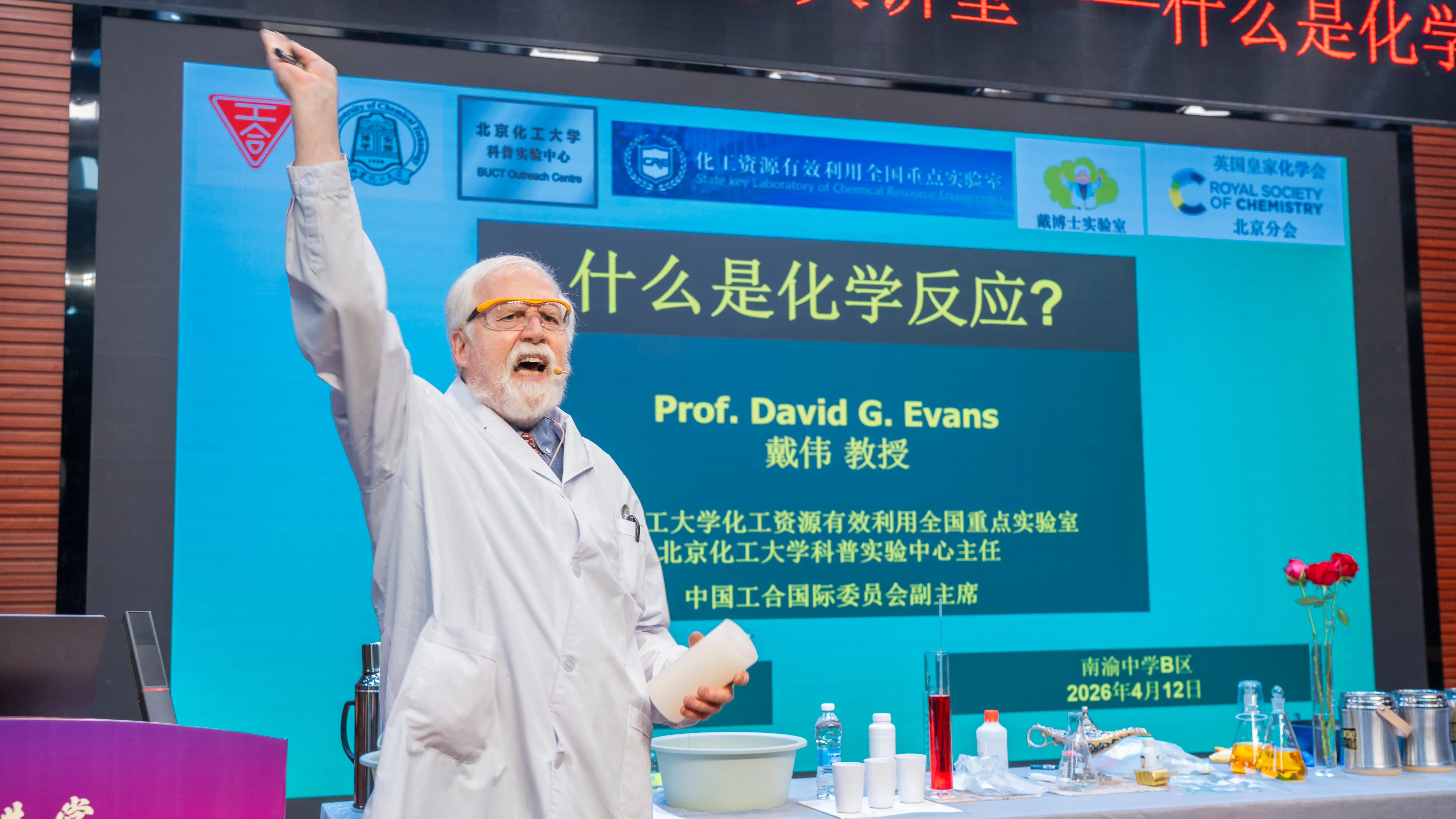 RSC in China - Professor David G. Evans Professor David G. Evans stands and shouts in a white coat holding a bottle and a white container, in front of a screen with his name on it in English and Chinese