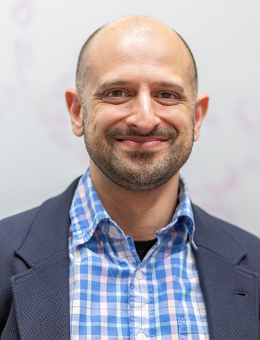 Headshot of Neal Mankad smiling, wearing blue chequered shirt and grey blazer