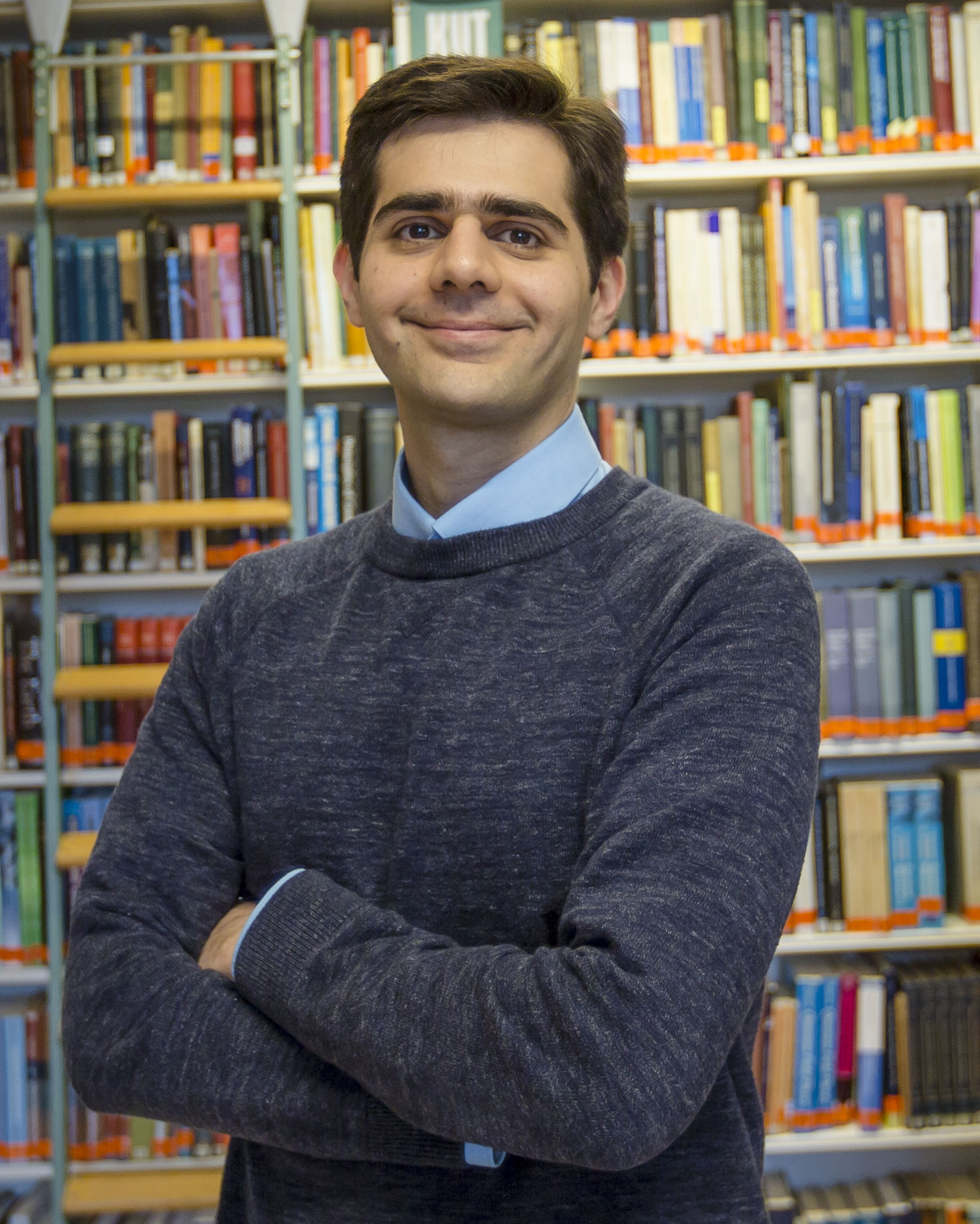 Amin Doostmohammadi smiling with his arms crossed, wearing a dark grey sweater in front of tall bookshelves.