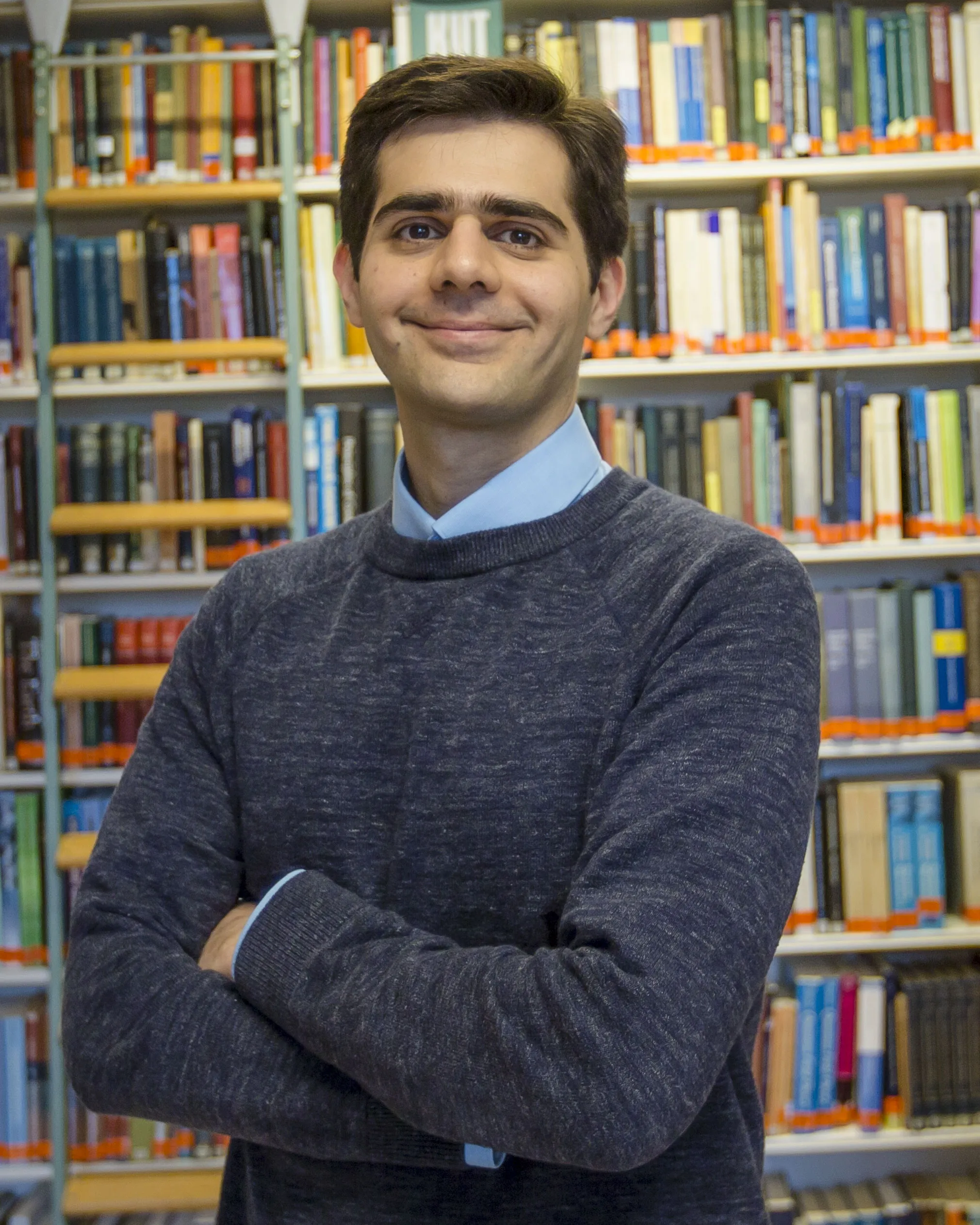 Amin Doostmohammadi Amin Doostmohammadi smiling with his arms crossed, wearing a dark grey sweater in front of tall bookshelves.