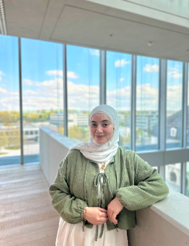 Rim Rihana wearing a light-colored hijab with green jacket, smiling while leaning against a balcony railing inside a modern building.