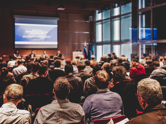 journal-lectureship-teaser-card A large audience attentively listens to a panel discussion during an in-person conference event