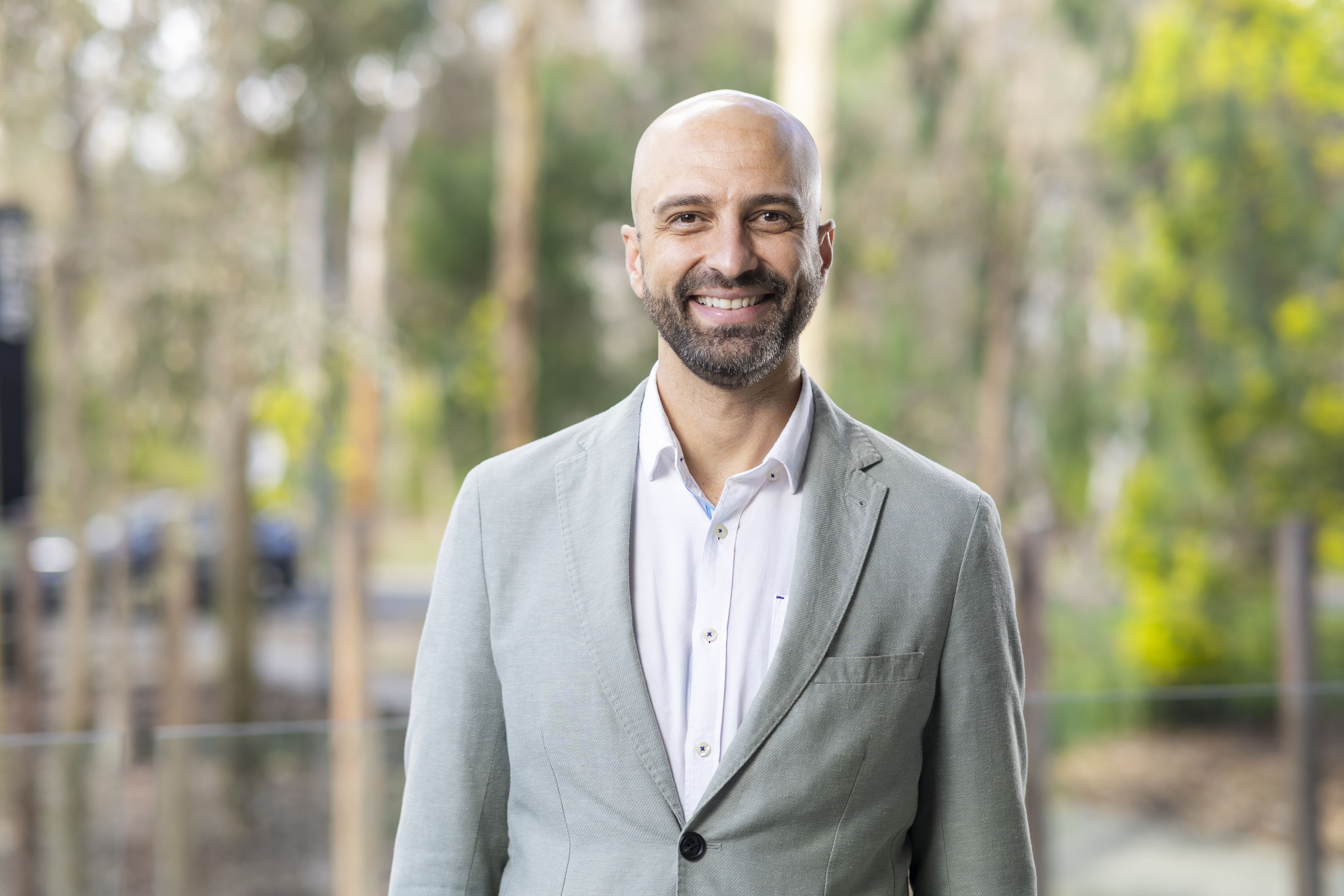 A close-up shot of Saimon Moraes Silva, wearing a light gray blazer against blurred background, showing out-of-focus green foliage.