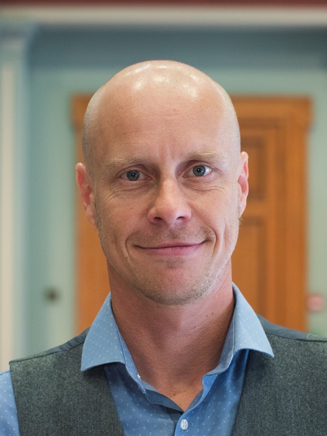Neil Hammond wearing a light blue shirt, with a softly blurred background showing a warm wooden door.