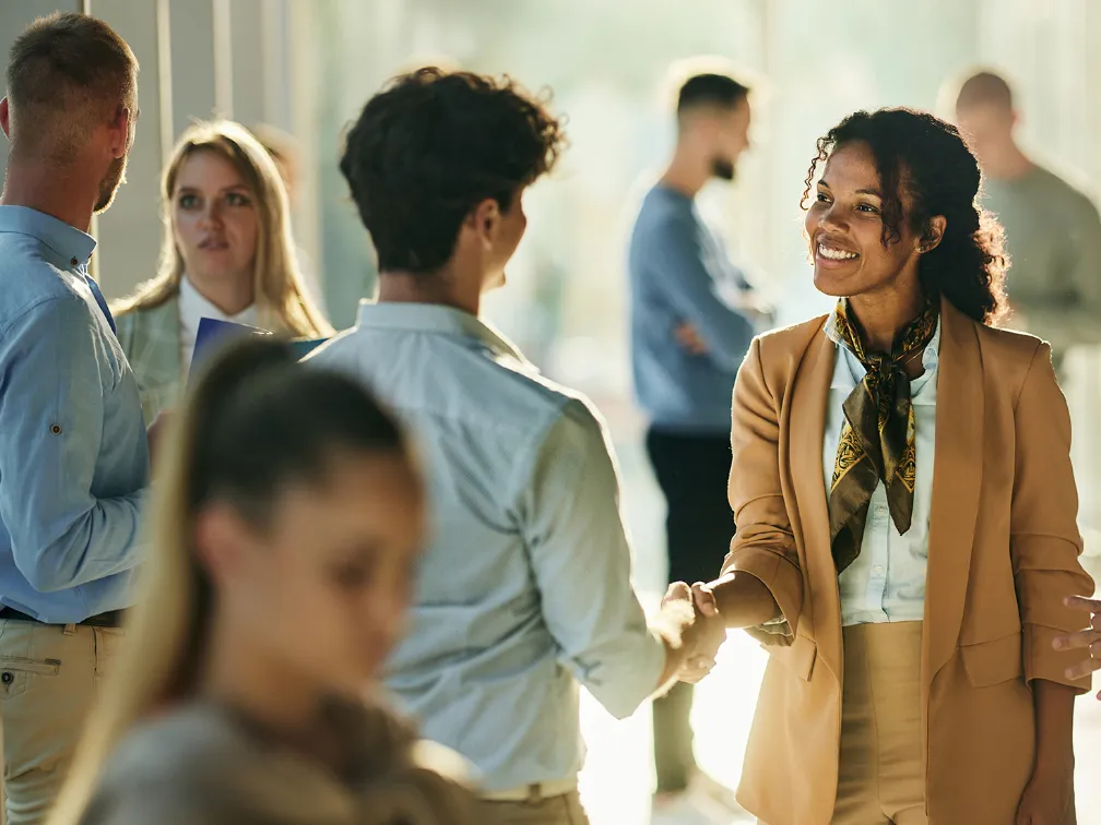 Candidate journey (image 1) A professional woman wearing a beige blazer over a white collared shirts miles warmly as she shakes hands with a man at a networking event.