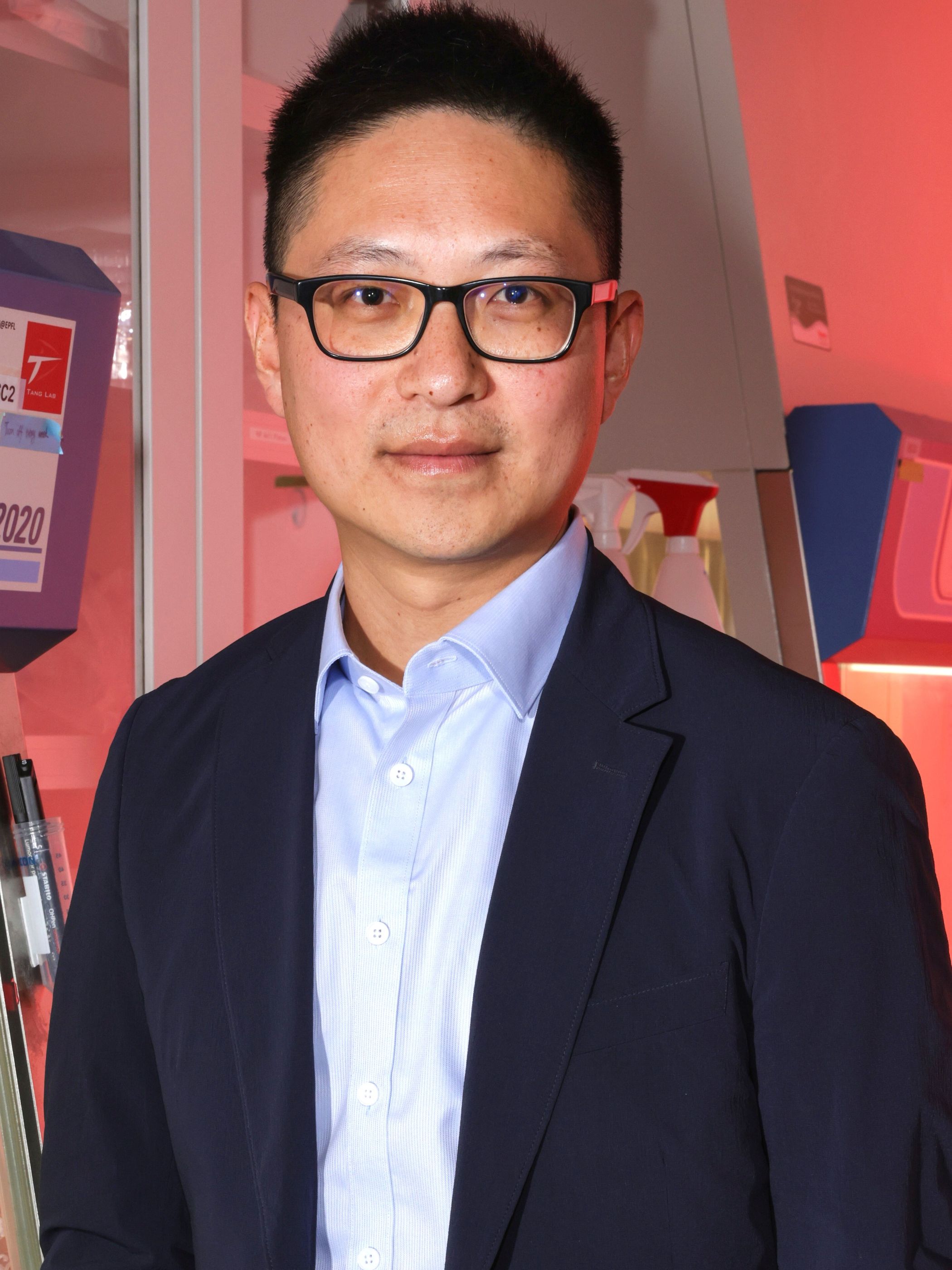 Li Tang facing camera wearing glasses and blue suit and open neck blue shirt