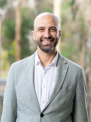 A close-up shot of Saimon Moraes Silva, wearing a light gray blazer against blurred background, showing out-of-focus green foliage.