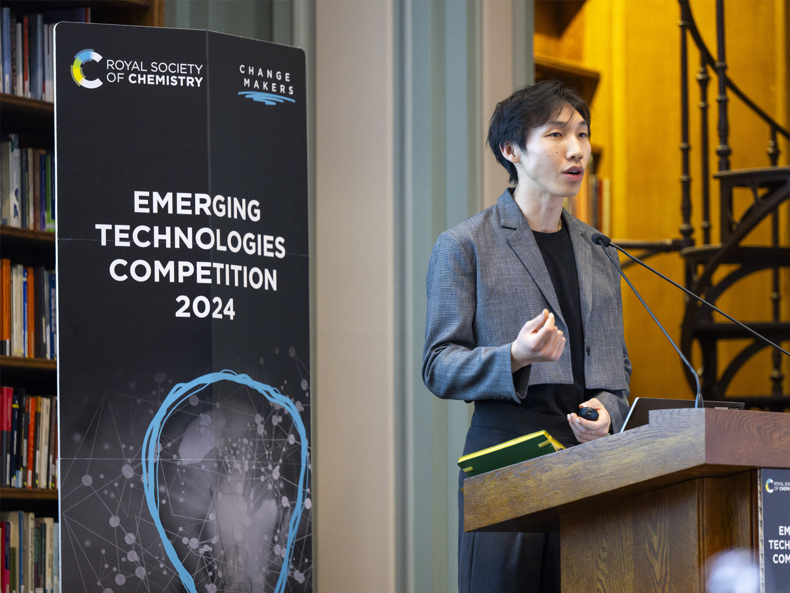 A male speaker talks into a microphone on a lectern with a standing banner to the left of him reading "Emerging Technologies Competition 2024'