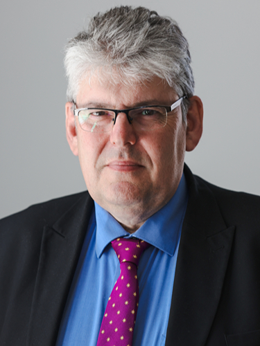 Paul Monks looking at the camera wearing a blue shirt, pink tie and black coat.