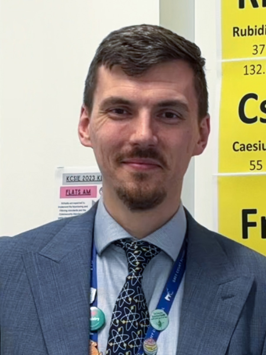 Matthew Gundry smiling to camera in suit and tie in front of periodic table