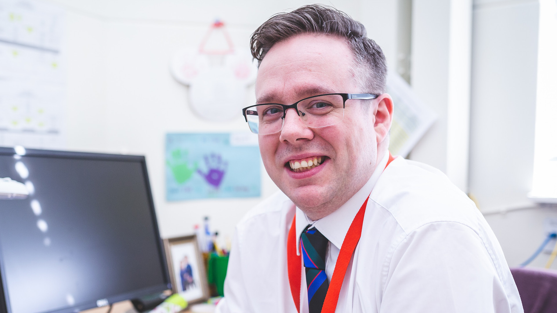 Kevin Morgan smiling to camera in glasses, white shirt and tie, at his desk
