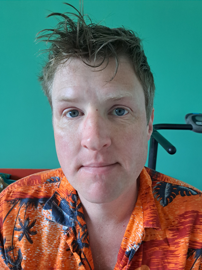 Michael Spencelayh wearing a vibrant orange shirt with patterns, sitting against a turquoise wall.
