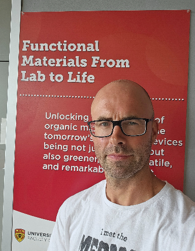 Gregory Welch wearing black-framed rectangular glasses and a white T-shirt against a red banner with white text that reads "Functional Materials From Lab to Life".